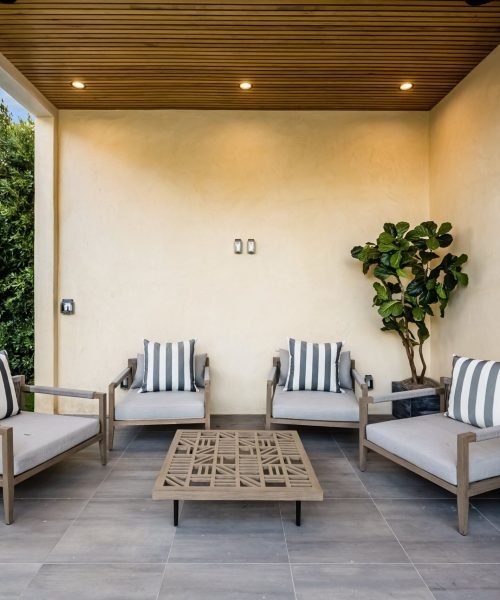 Modern outdoor patio with wooden ceiling, cushioned chairs, striped pillows, and a coffee table, surrounded by greenery.